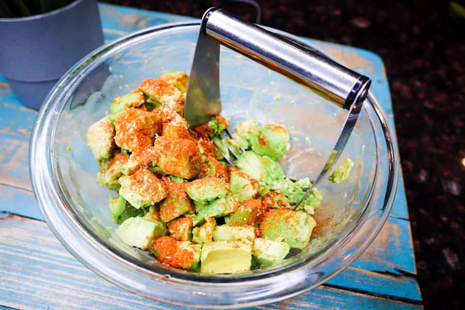 Avocado and spices in a glass bowl with a pastry blender.