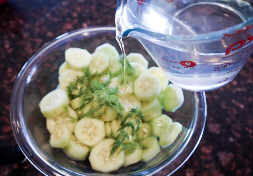 Brine being poured over cucumber slices and dill in a bowl for Super Simple Refrigerator Pickles.