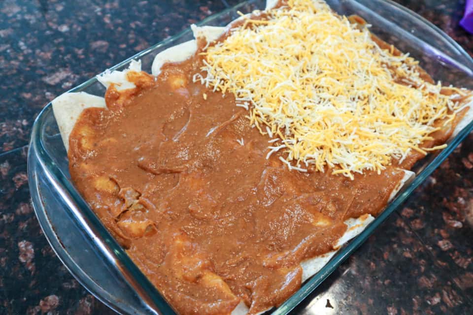 Picture of enchilada sauce and cheese going on top of rolled tortillas for Baked Chicken Enchiladas.