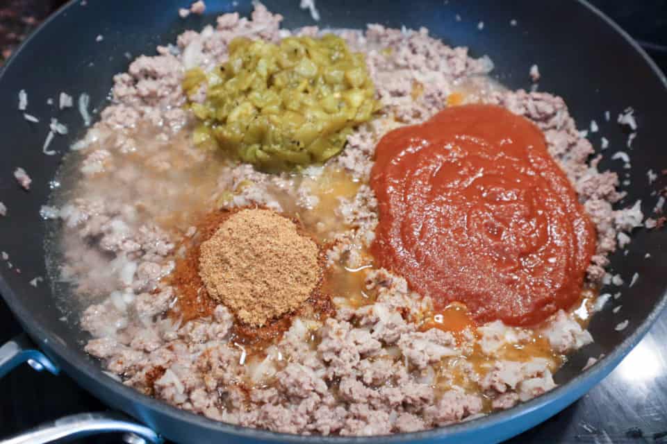 Taco seasoning, taco sauce and green chilis being added to the ground beef mixture in the skillet for Oven Baked Taco Casserole.