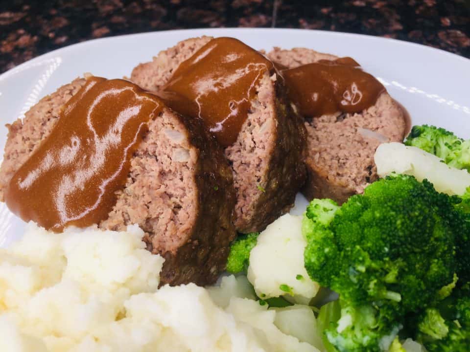 Finished Simple Weeknight Meatloaf, sliced with gravy, on a plate with potatoes and broccoli.
