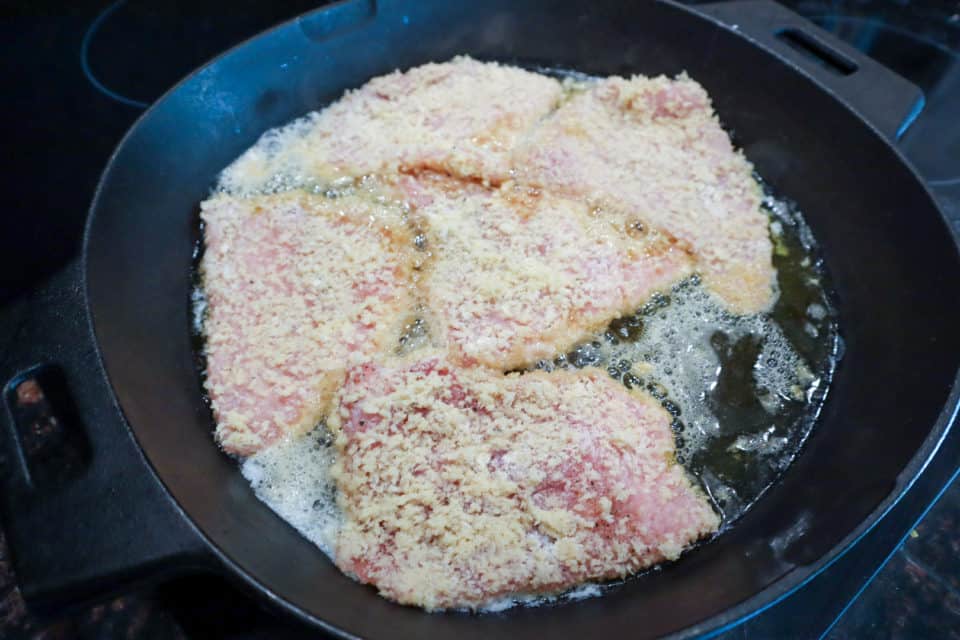 Breaded pork chops frying in a cast iron pan for Pork Scallopini with Lemon Caper Sauce.
