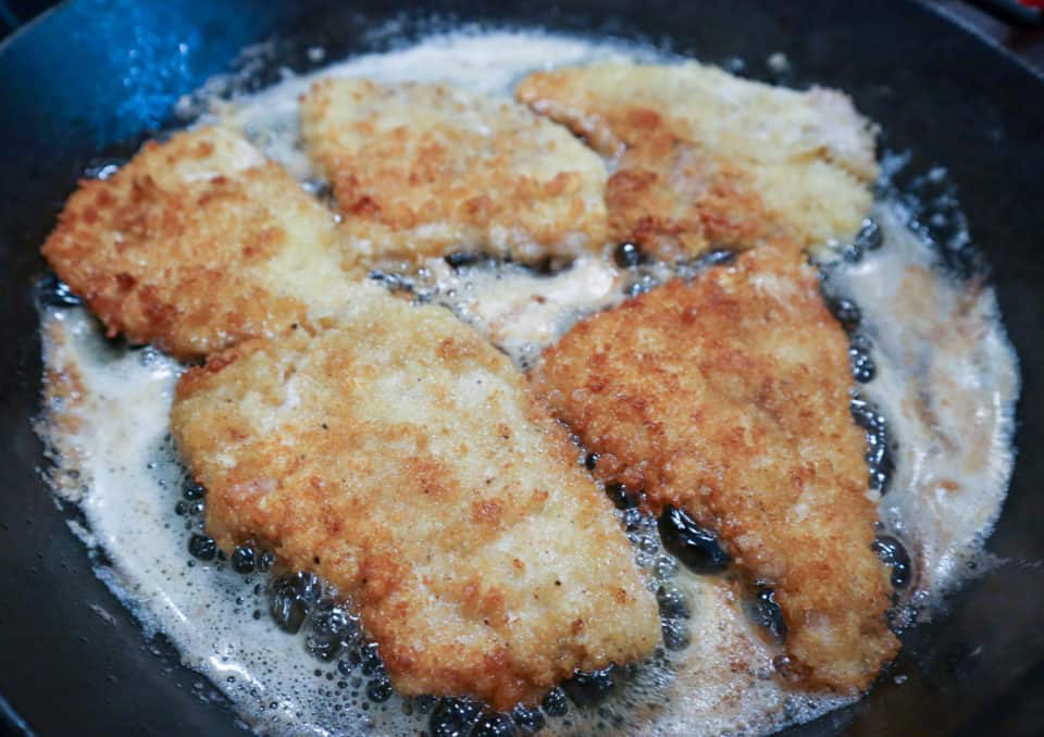 Breaded pork chops frying in a cast iron pan for Pork Scallopini with Lemon Caper Sauce.