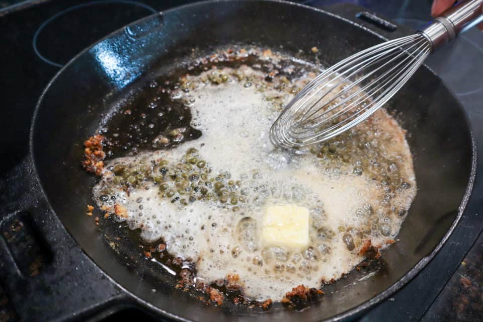 Capers and butter melting in a cast iron pan, brown bits being scraped loose from the pan with a whisk.