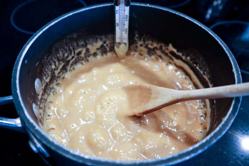 Bubbling Homemade Soft Caramels ingredients with a candy thermometer and a wooden spoon in a saucepan.