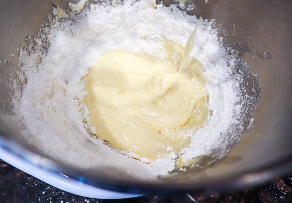 Powdered confectioners sugar being mixed into butter mixture in a bowl.