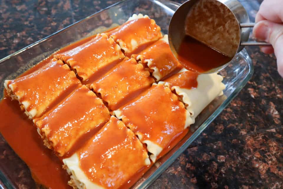 Rolled noodles filled with enchilada filling in the baking dish being covered in enchilada sauce.