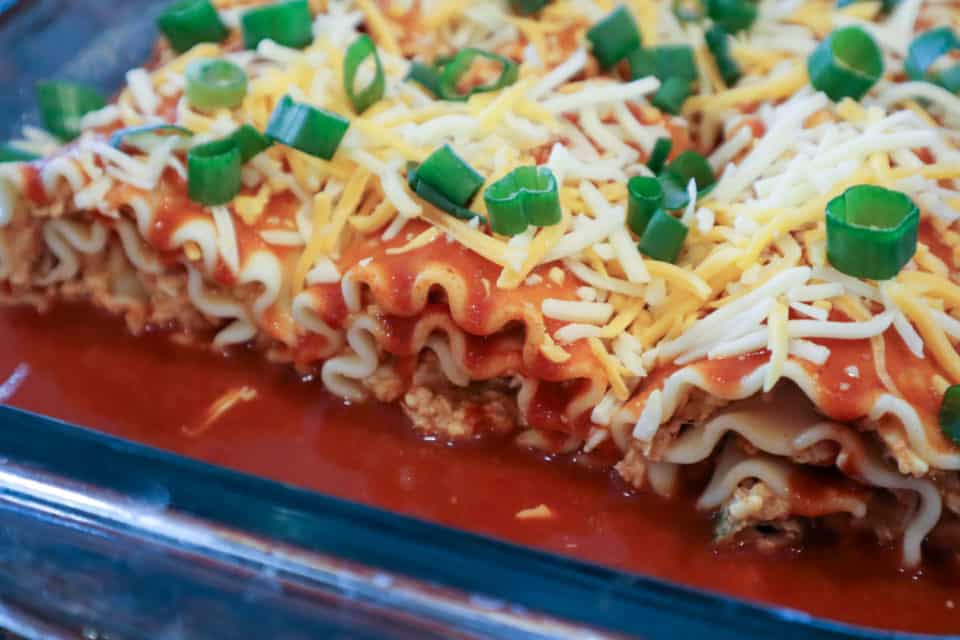 Rolled noodles filled with enchilada filling in the baking dish covered with enchilada sauce, shredded cheese and green onions.