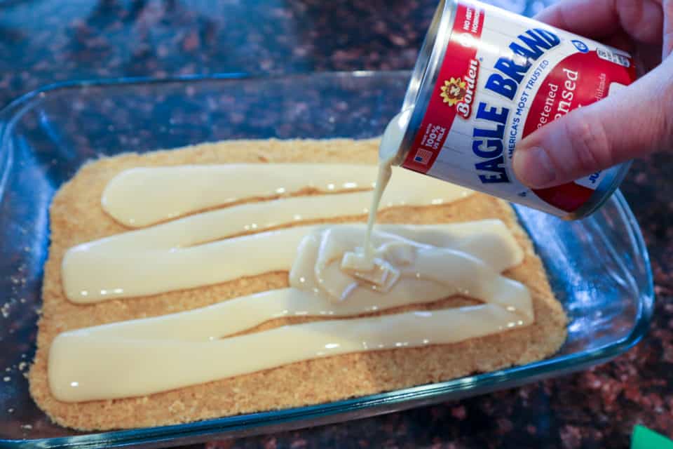 Pouring sweetened condensed milk over the graham cracker crust layer.