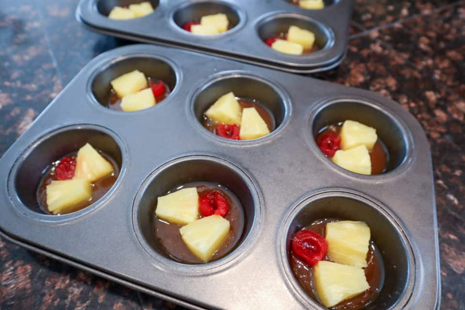 Muffin tin with brown sugar sauce, pineapple chunks and a maraschino cherry.