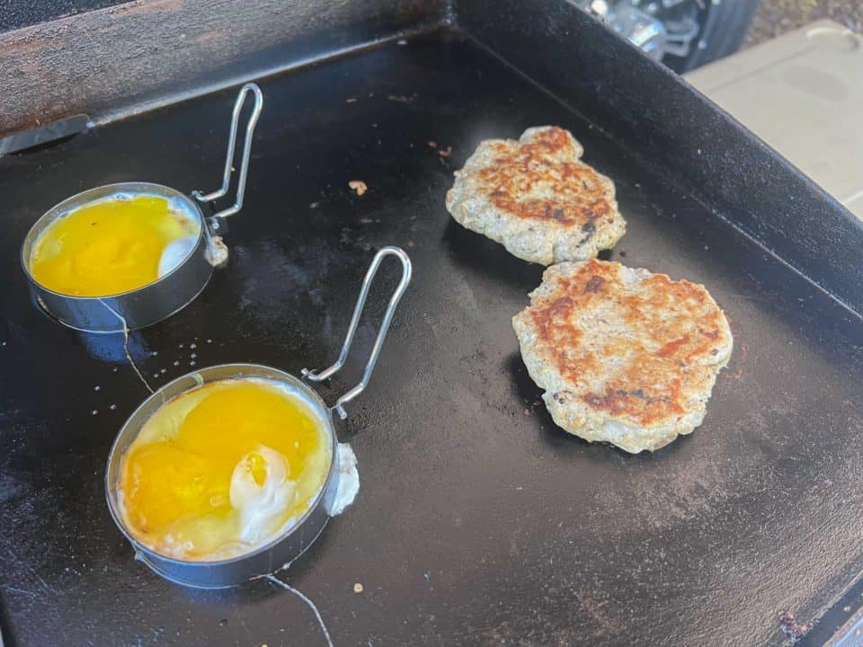 Cooking eggs in rings on the griddle beside the cooked Ground Turkey Breakfast Sausage for breakfast sandwiches.
